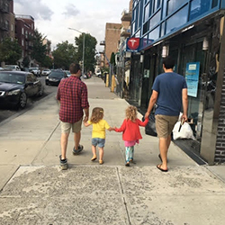 Two dads and two daughters all walking hand in hand down an open sidewalk