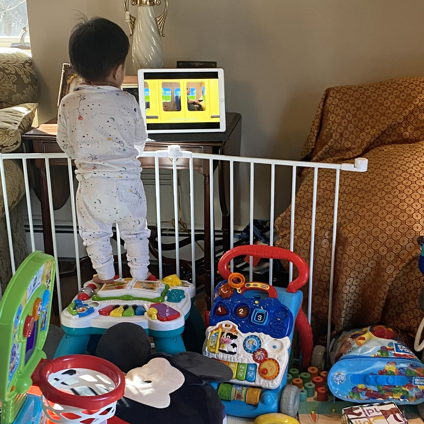 Baby wearing pajamas and standing on top of some baby toys while watching an iPad video