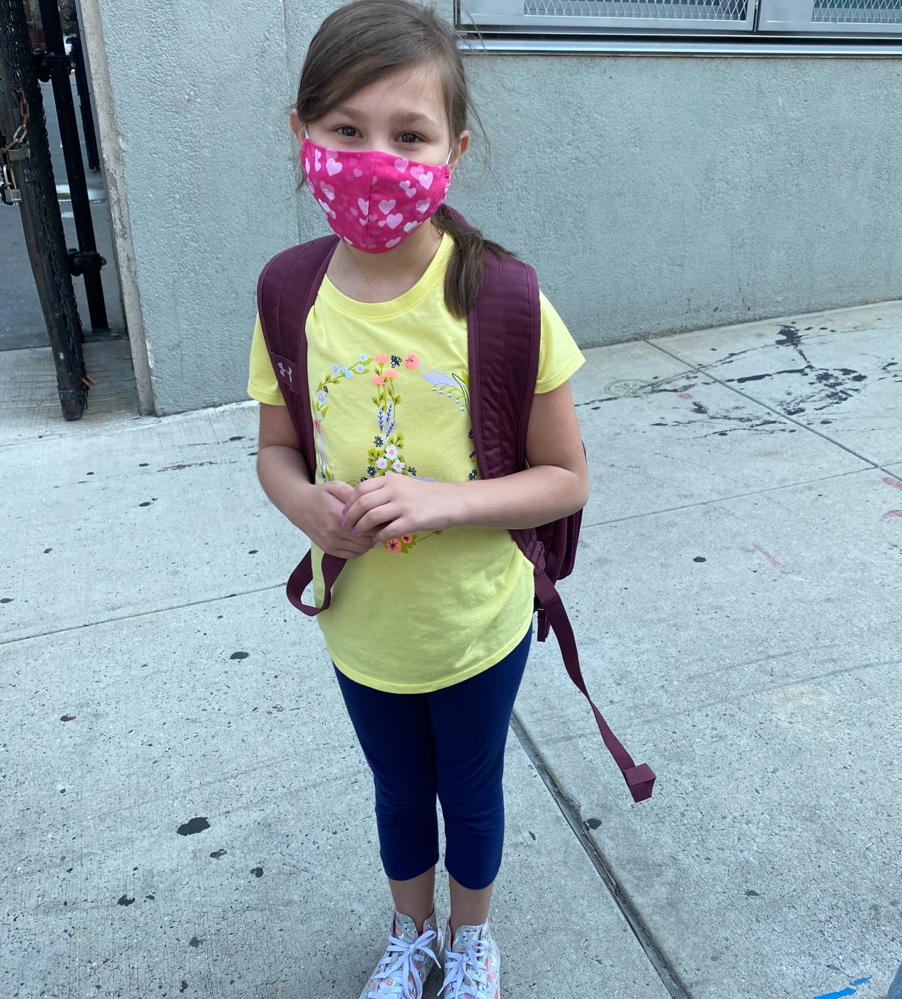 Young girl wearing a backpack and a fabric face mask outside of a school