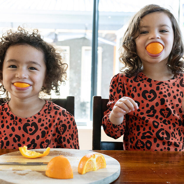 Two young girls sitting at a table wearing matching pajamas and eating oranges